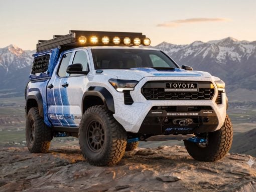 White Toyota pickup with roof rack and eight lights, parked on a rocky overlook with snow-capped mountains in the background.