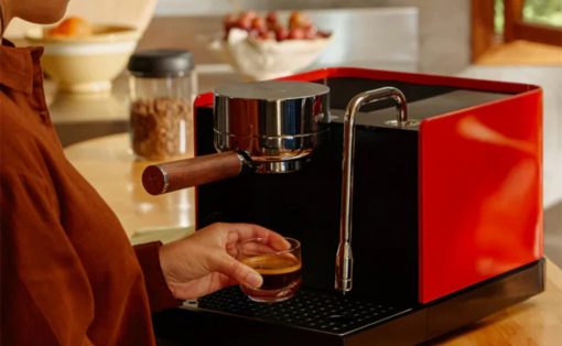 Person using a red and black home espresso machine, holding a small glass of espresso under the spout.