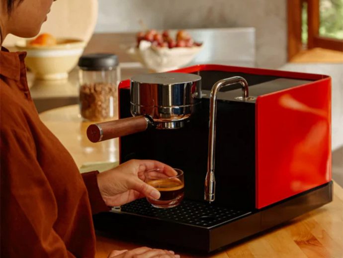 Person using a red and black home espresso machine, holding a small glass of espresso under the spout.