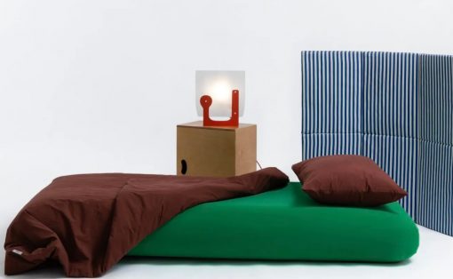 Minimalist bed setup: green mattress with brown duvet and pillow, blue striped headboard, and a wooden nightstand with a red abstract lamp.
