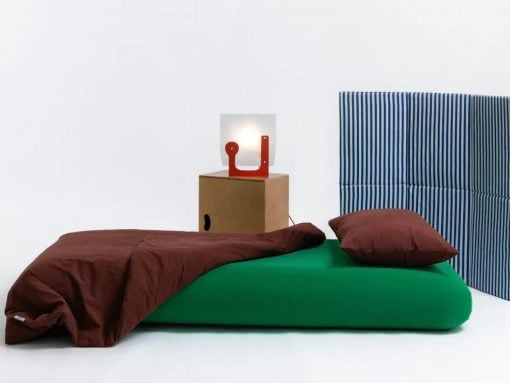 Minimalist bed setup: green mattress with brown duvet and pillow, blue striped headboard, and a wooden nightstand with a red abstract lamp.