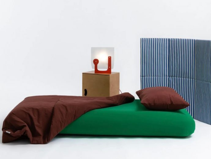 Minimalist bed setup: green mattress with brown duvet and pillow, blue striped headboard, and a wooden nightstand with a red abstract lamp.