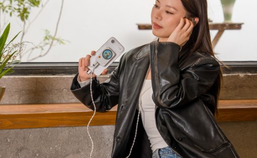 Woman in a black leather jacket sits at a wooden counter, holding a white handheld device with a small screen and cord, looking to the side.