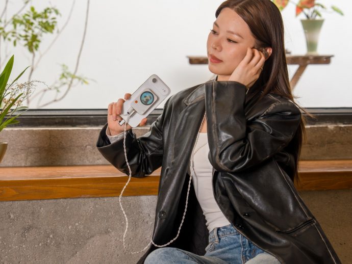 Woman in a black leather jacket sits at a wooden counter, holding a white handheld device with a small screen and cord, looking to the side.