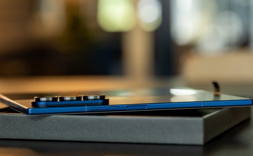 Blue smartphone with a large multi-lens camera module resting on a gray box on a dark desk, background softly blurred.