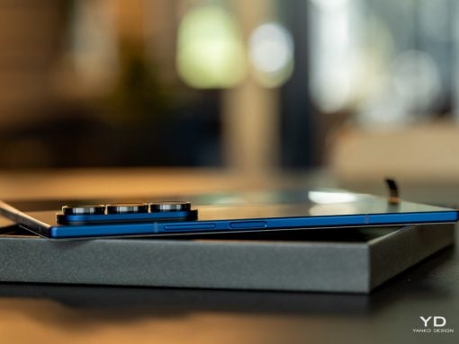 Blue smartphone with a large multi-lens camera module resting on a gray box on a dark desk, background softly blurred.