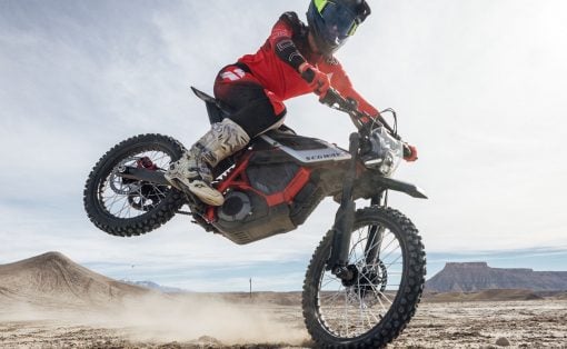 Rider in red gear and full-face helmet jumps a Segway off-road bike over dusty desert terrain with distant mesas in view.
