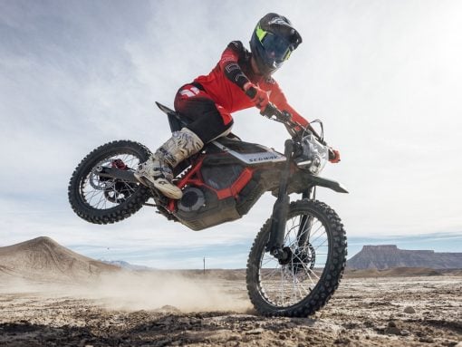 Rider in red gear and full-face helmet jumps a Segway off-road bike over dusty desert terrain with distant mesas in view.