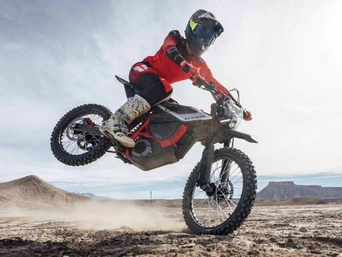 Rider in red gear and full-face helmet jumps a Segway off-road bike over dusty desert terrain with distant mesas in view.