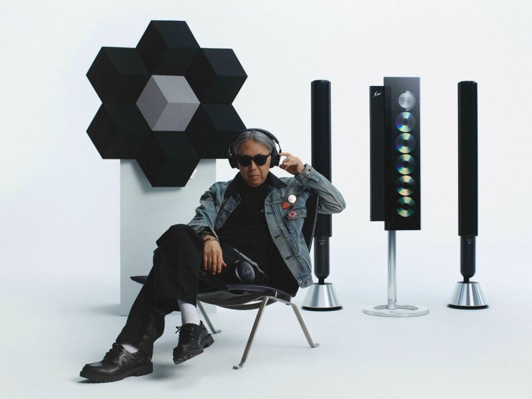 Person wearing sunglasses and a denim jacket sits in a studio with a modular black hexagon sculpture and tall speaker cabinets in the background.