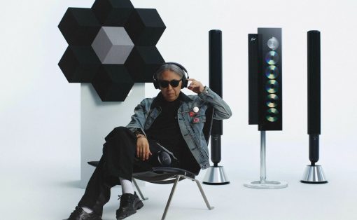 Person wearing sunglasses and a denim jacket sits in a studio with a modular black hexagon sculpture and tall speaker cabinets in the background.