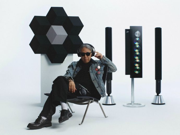 Person wearing sunglasses and a denim jacket sits in a studio with a modular black hexagon sculpture and tall speaker cabinets in the background.