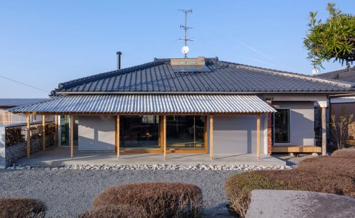 House with blue ceramic tiled roof, wooden porch posts, and a gravel front yard. Large glass doors run along the front of the home with a covered porch area.