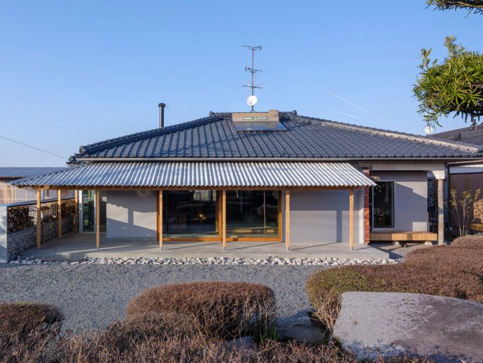 House with blue ceramic tiled roof, wooden porch posts, and a gravel front yard. Large glass doors run along the front of the home with a covered porch area.