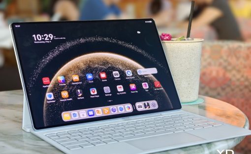 Tablet on a light keyboard case displaying a home screen with app icons, placed on a marble table in a cafe beside a speckled cup with a straw