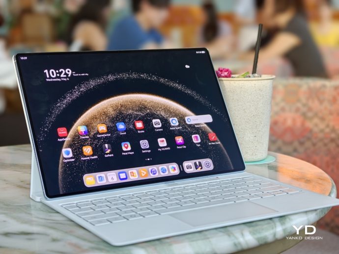 Tablet on a light keyboard case displaying a home screen with app icons, placed on a marble table in a cafe beside a speckled cup with a straw