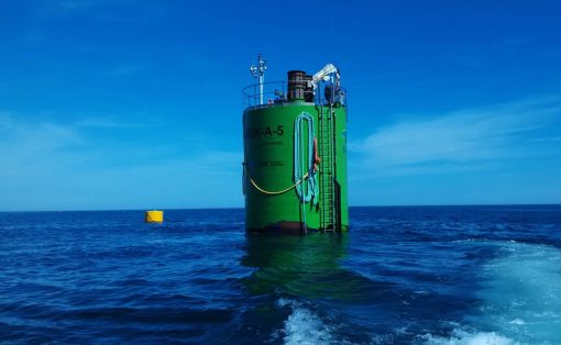 Large green cylindrical offshore structure with a ladder and safety railing rising from the blue sea under a clear sky, with coiled rope along its side and a distant yellow buoy nearby