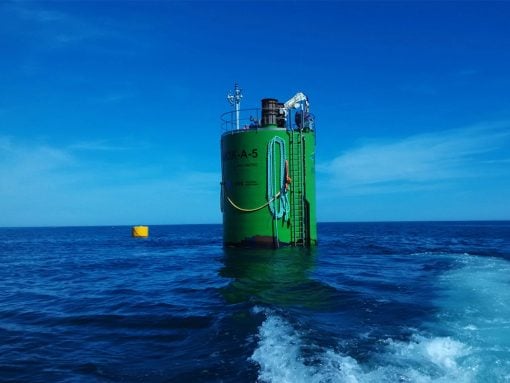 Large green cylindrical offshore structure with a ladder and safety railing rising from the blue sea under a clear sky, with coiled rope along its side and a distant yellow buoy nearby