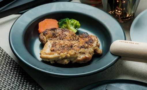 Juicy, pepper-seasoned chicken leg on a blue plate with broccoli and carrot; wooden-handled frying pan in view.