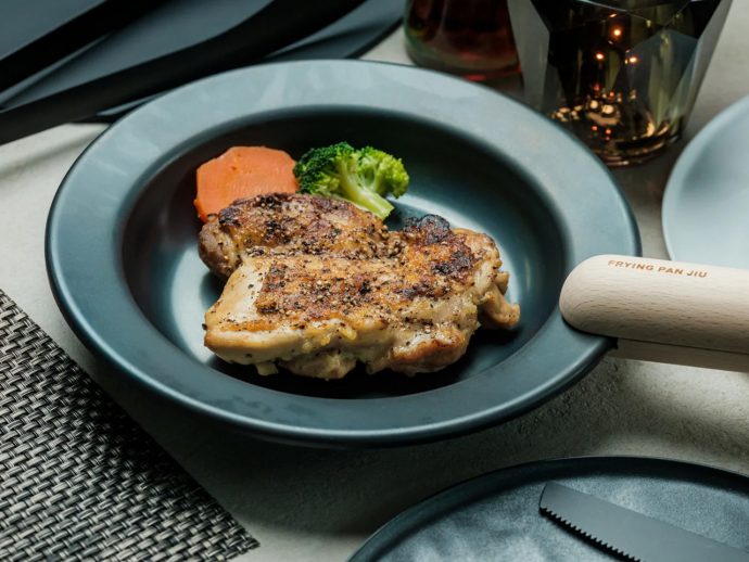 Juicy, pepper-seasoned chicken leg on a blue plate with broccoli and carrot; wooden-handled frying pan in view.