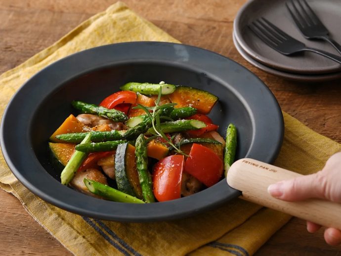 Pan with sautéed vegetables (asparagus, bell pepper, zucchini, squash) and chicken, garnished with rosemary, on a dark skillet with a wooden handle.