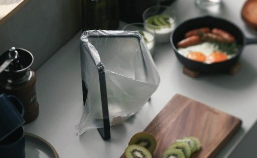 Tall blender wrapped in plastic on a kitchen counter, with kiwi slices on a wooden cutting board and a skillet of eggs and bacon in the background.