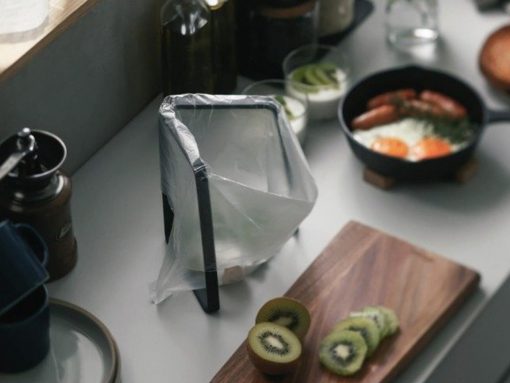 Tall blender wrapped in plastic on a kitchen counter, with kiwi slices on a wooden cutting board and a skillet of eggs and bacon in the background.