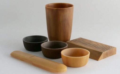 Collection of wooden kitchenware: tall cup, several bowls in black and natural wood tones, a rectangular cutting board, and a long spatula on a light surface.