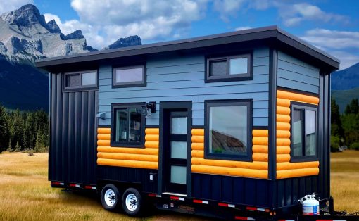 Tiny house on a trailer parked in a grassy field with mountains in the background, blue siding and orange log accents visible on the exterior walls