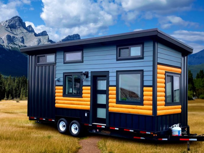 Tiny house on a trailer parked in a grassy field with mountains in the background, blue siding and orange log accents visible on the exterior walls
