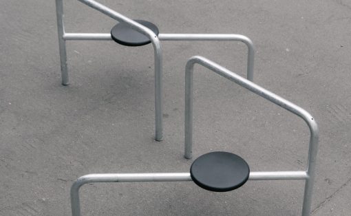 Outdoor playground fitness equipment: three silver metal bars forming low arches with black circular seats anchored to the ground on a paved surface