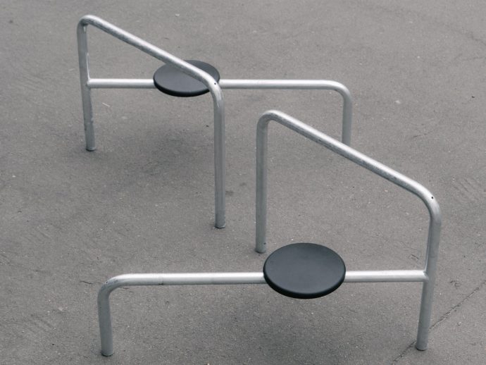 Outdoor playground fitness equipment: three silver metal bars forming low arches with black circular seats anchored to the ground on a paved surface