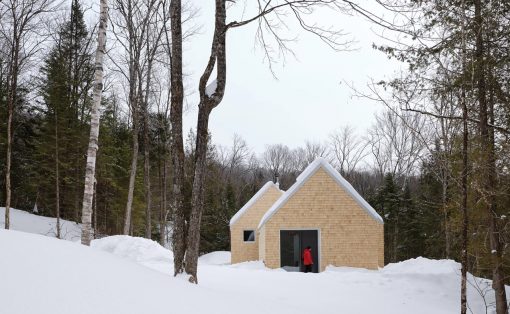Small wooden cabin in a snowy forest with a person in a red jacket entering through the slider door.