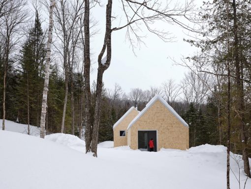 Small wooden cabin in a snowy forest with a person in a red jacket entering through the slider door.