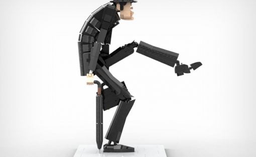 LEGO minifigure in dark armor and helmet stands on a white display base, bending forward and reaching outward with a long black tool-like piece in one hand.