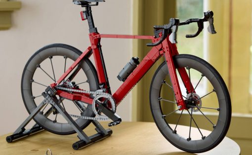 Red LEGO road bicycle mounted on a trainer stand indoors, with a gray water bottle on the frame and keys on the table nearby.