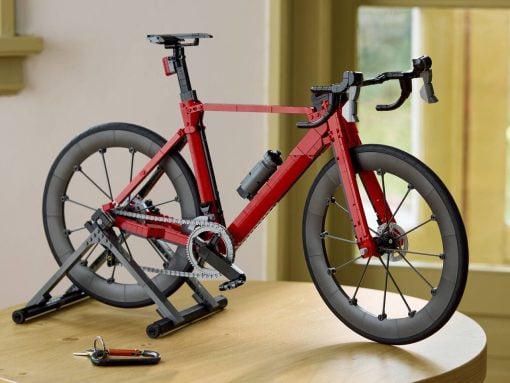 Red LEGO road bicycle mounted on a trainer stand indoors, with a gray water bottle on the frame and keys on the table nearby.