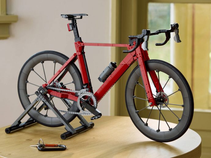 Red LEGO road bicycle mounted on a trainer stand indoors, with a gray water bottle on the frame and keys on the table nearby.