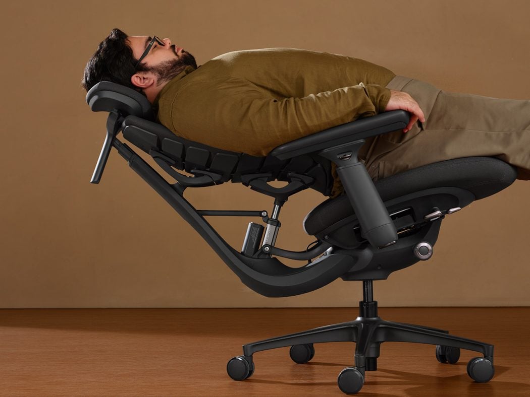 Man reclining in a black ergonomic lounge chair with a padded headrest and wheeled base, resting on a wooden floor.