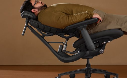 Man reclining in a black ergonomic lounge chair with a padded headrest and wheeled base, resting on a wooden floor.