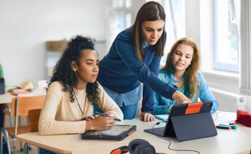 Three students work on tablets at a bright classroom table while a teacher guides them.