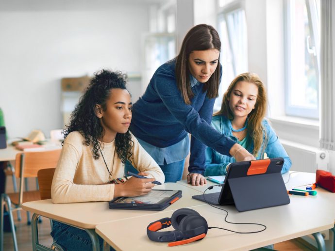 Three students work on tablets at a bright classroom table while a teacher guides them.
