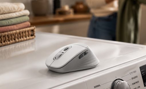 White wireless computer mouse resting on a washing machine, with folded towels and a blurred laundry area in the background.
