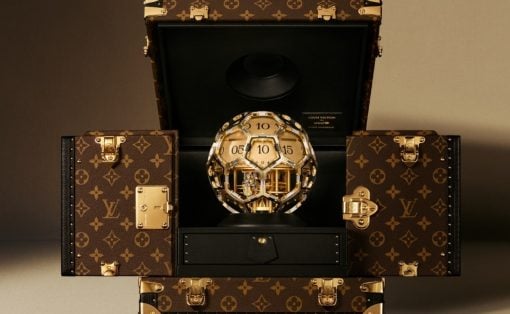 Gold, faceted spherical timepiece displayed inside an open Louis Vuitton trunk, surrounded by LV monogram luggage cases.