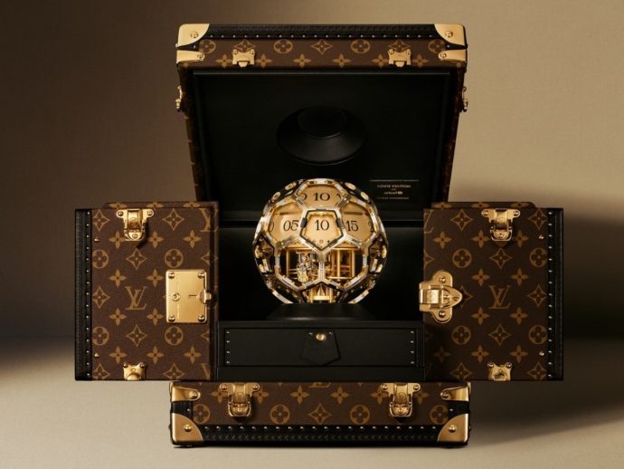 Gold, faceted spherical timepiece displayed inside an open Louis Vuitton trunk, surrounded by LV monogram luggage cases.