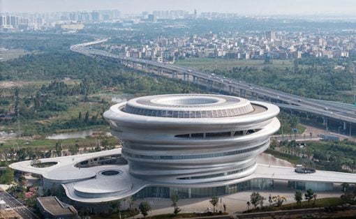 Futuristic circular white building with stacked rings beside a highway and green fields, city in the distance.