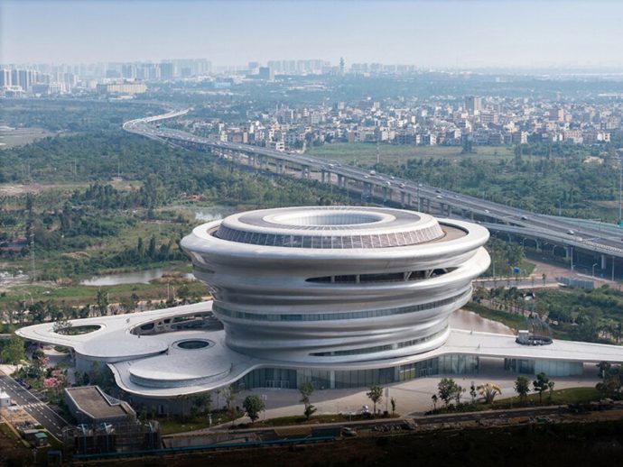 Futuristic circular white building with stacked rings beside a highway and green fields, city in the distance.