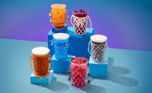 Colorful drink cups wrapped in beaded jewelry displayed on blue stepped platforms against a gradient background.