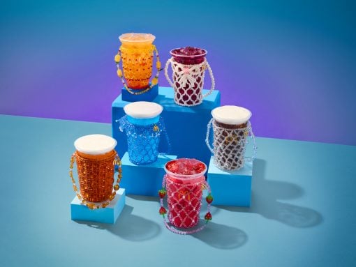 Colorful drink cups wrapped in beaded jewelry displayed on blue stepped platforms against a gradient background.