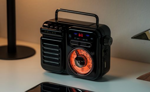 Black retro portable radio with orange tuning dial and handle on a desk beside a smartphone reflection.